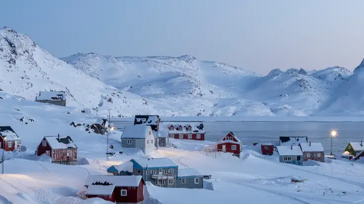 Et billede af en bygd i Grønland ved sneklædte bakker og vand.