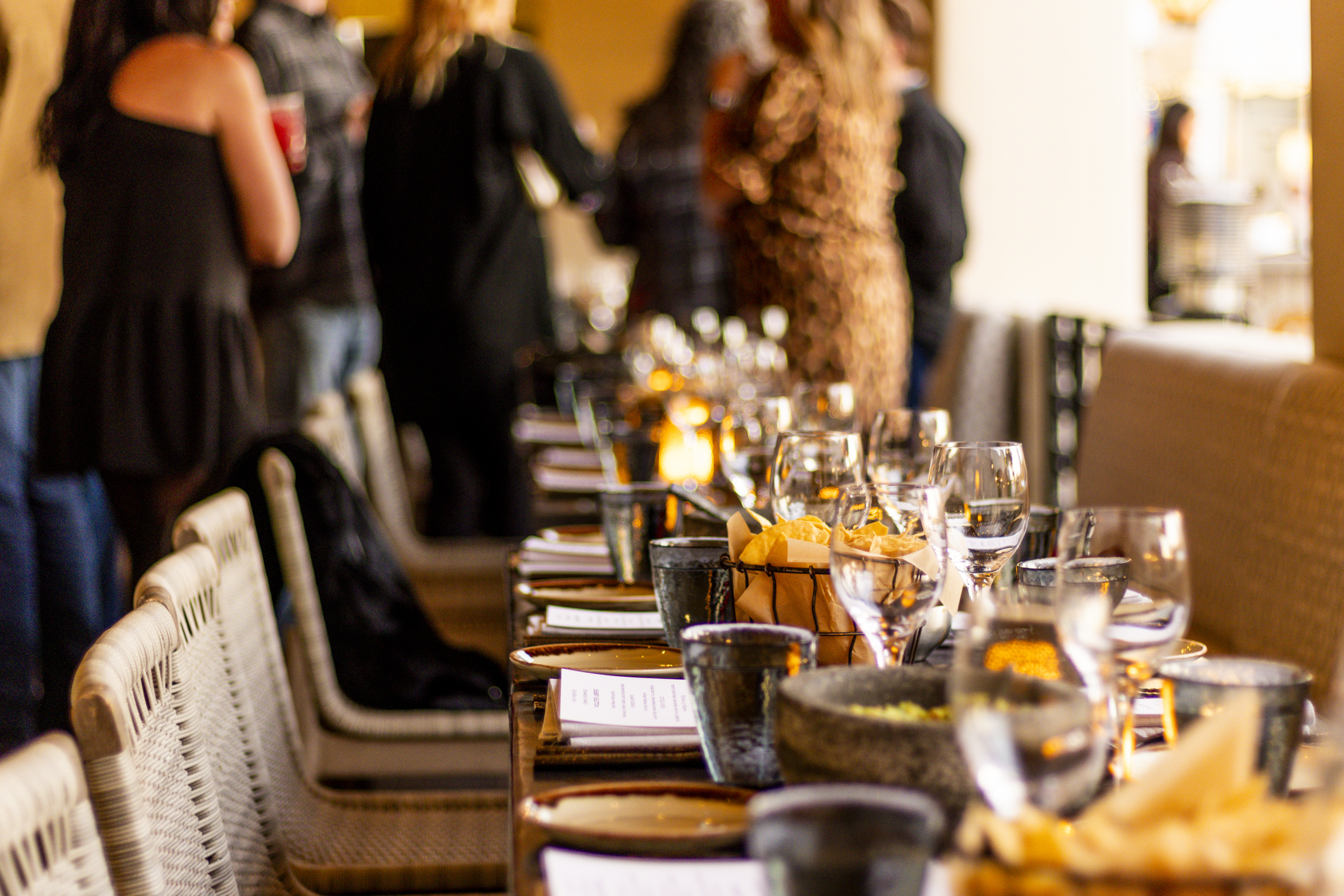 Langt dækket middagsbord med glas, service og levende lys i varmt lys, mens gæster står og mingler i baggrunden i en hyggelig og elegant restaurantstemning.