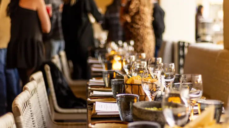 Langt dækket middagsbord med glas, service og levende lys i varmt lys, mens gæster står og mingler i baggrunden i en hyggelig og elegant restaurantstemning.