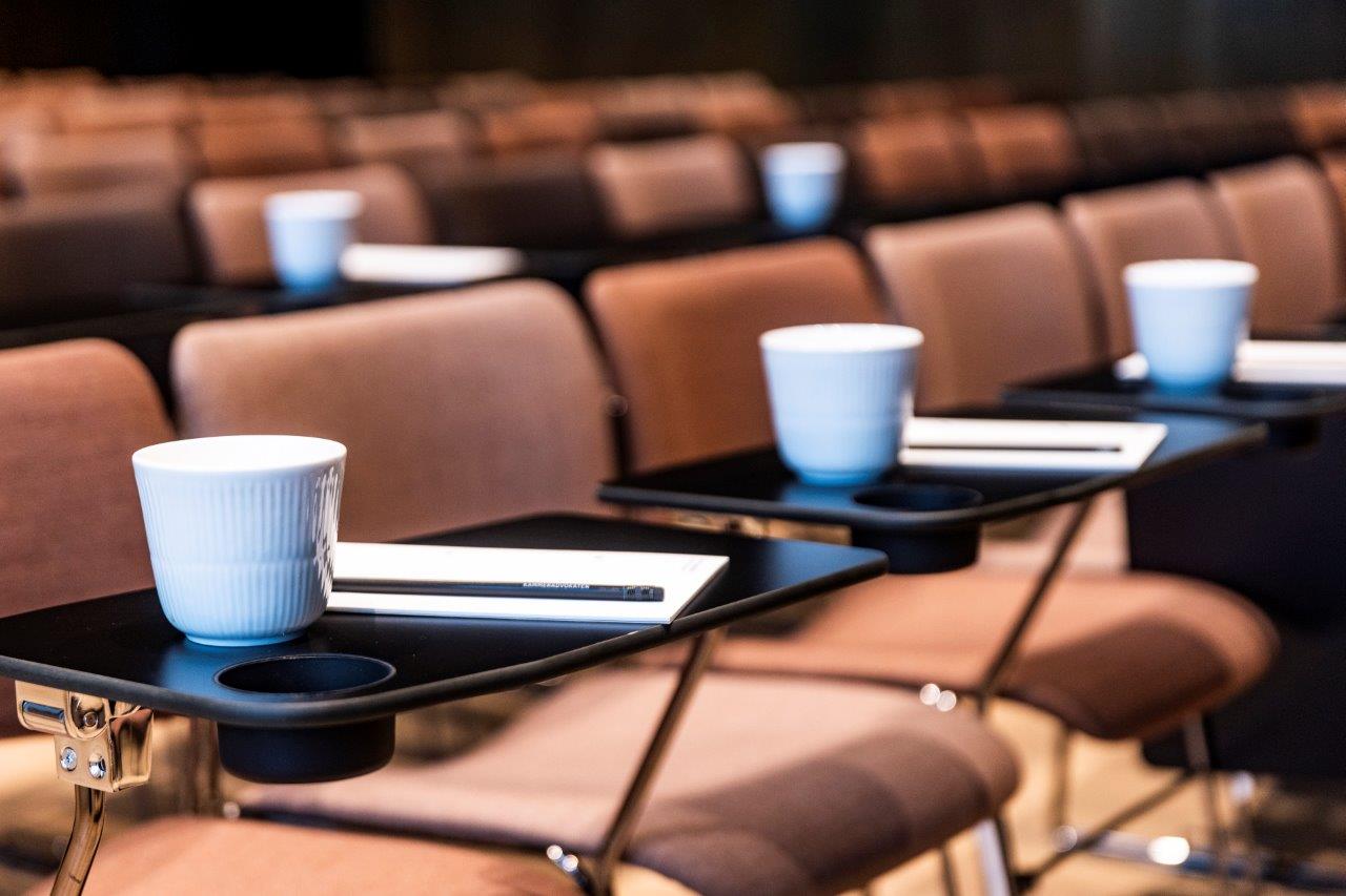 Nærbillede af pænt opstillede konferencestole med kaffekrus og notesblokke på små bakker, klar til deltagere ved et formelt arrangement eller seminar.