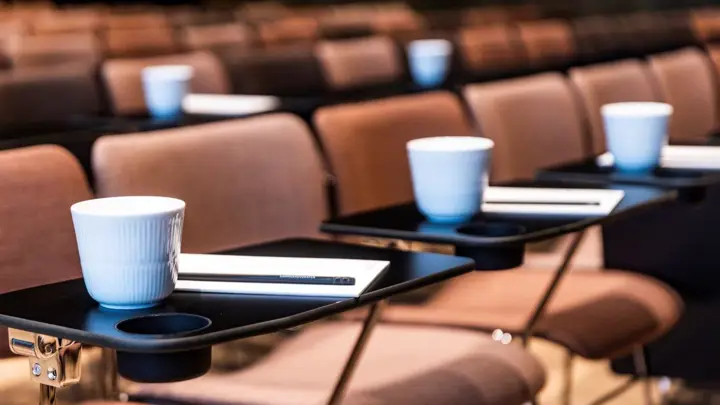 Nærbillede af pænt opstillede konferencestole med kaffekrus og notesblokke på små bakker, klar til deltagere ved et formelt arrangement eller seminar.