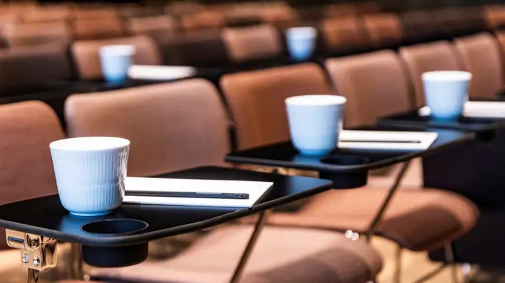 Nærbillede af pænt opstillede konferencestole med kaffekrus og notesblokke på små bakker, klar til deltagere ved et formelt arrangement eller seminar.