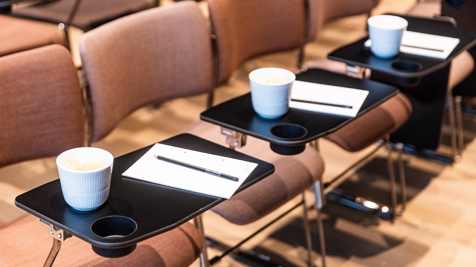Stole i et auditorium med notesblokke, blyanter og kaffekoppe på bordene foran dem.