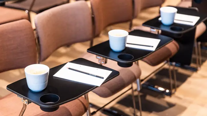 Stole i et auditorium med notesblokke, blyanter og kaffekoppe på bordene foran dem.